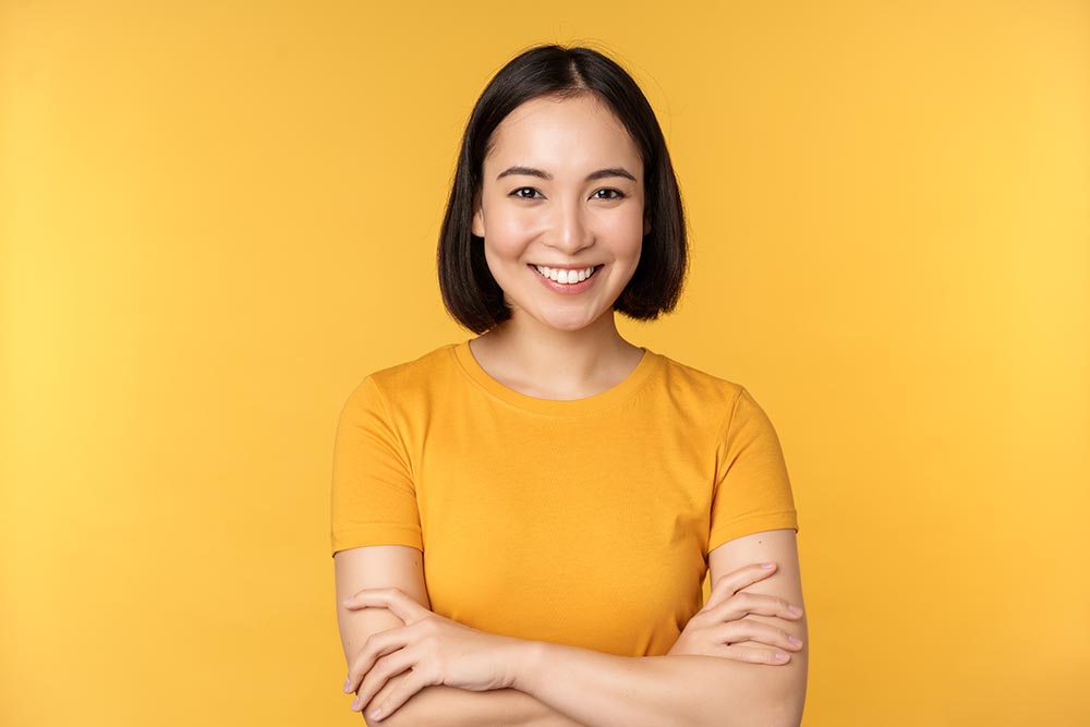 Confident asian girl cross arms on chest, smiling and looking assertive, standing over yellow background