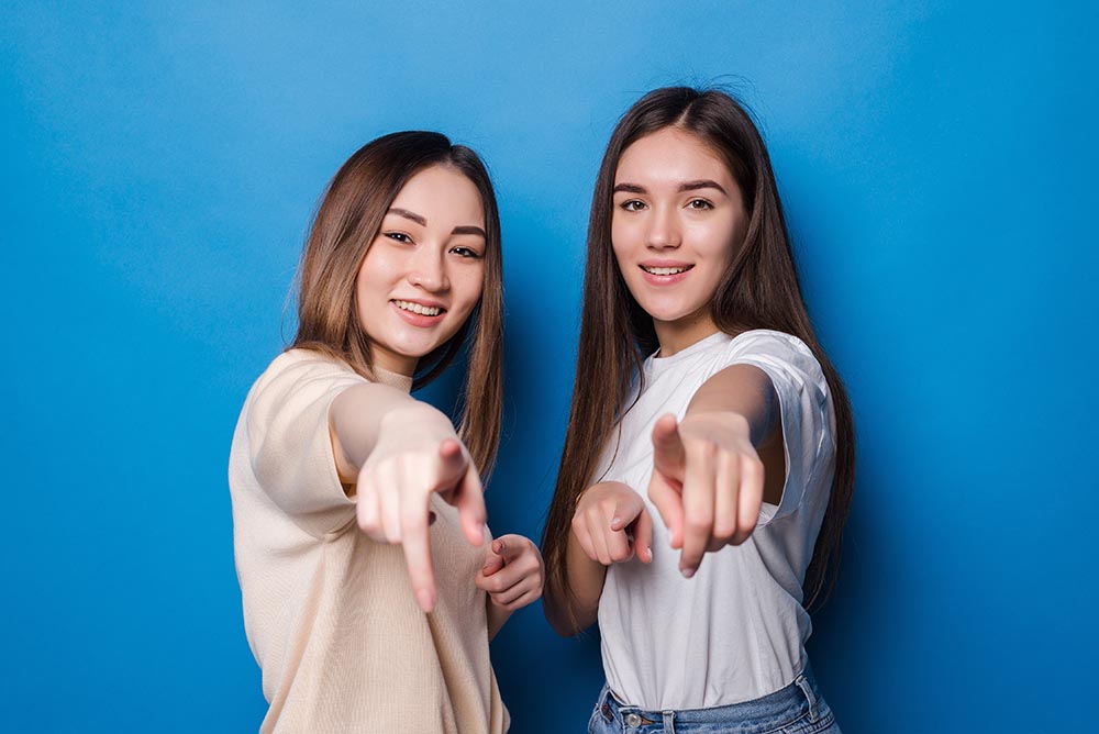Two cheerful elegant girls smiling and pointing fingers at camera meaning hey you isolated over blue background